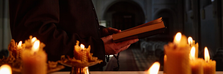 Male priest standing in church holding book with both hands, reading near burning candles, wearing...