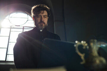 Caucasian middle aged man with beard wearing clerical collar standing in church near window reading...