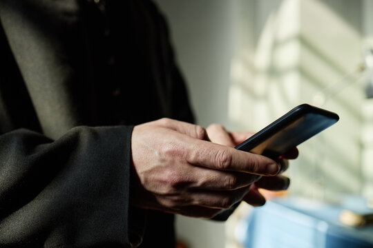 Man holding smartphone in hands, standing in church, wearing religious robe, using mobile device for communication or reading, sunlight casting shadows