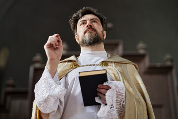 Caucasian middle aged man wearing priest vestments standing in church holding book and raising hand...