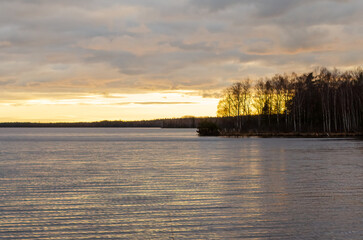 Calm sunset glow reflected on lake water with bare trees and dramatic clouds. Tranquil sunset scene, soft golden reflections on rippling water, low angle, lakeside serenity, peaceful atmosphere, moody
