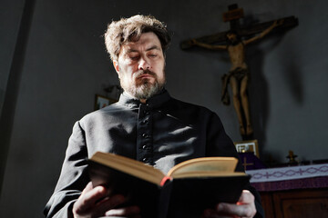 Caucasian middle aged man with beard wearing clerical robe standing in church reading book with...