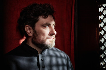 Middle aged Caucasian man with beard sitting in church wearing clerical clothing, gazing thoughtfully toward window with patterned light casting shadows on face and background