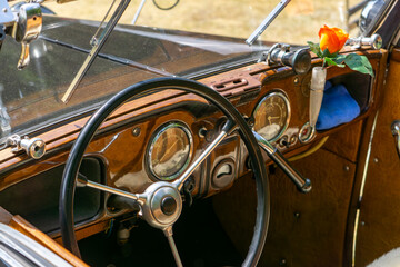 Wooden dashboard of a vintage car with a steering wheel, analog gauges, and a yellow rose in a small vase – stylish interior of a classic automobile.