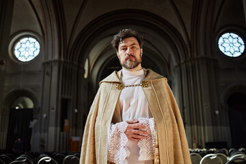 Portrait of middle aged man standing in church wearing ceremonial robe, hands clasped in front,...