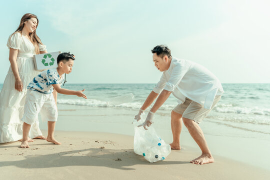 The son of an Asian family is throwing plastic bottles picked up from the beach in a garbage bag that his father is carrying for recycling or reuse. Dirty beach