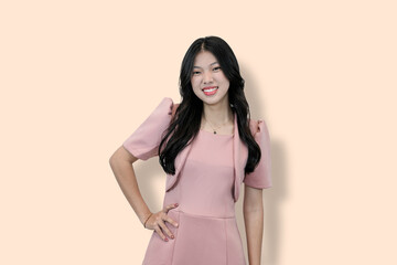 A young Asian woman in a pink dress and bolero smiles confidently at the camera, hand on hip,...