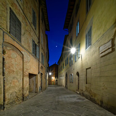 A narrow street in Siena, Italy, illuminated by warm streetlights under a dark blue night sky. The historic architecture, stone-paved path, and serene ambiance create a picturesque nighttime scene.