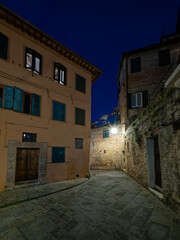 A small piazza in Siena, Italy, illuminated by warm streetlights under a dark blue night sky. The historic architecture, stone-paved square, and serene ambiance create a picturesque nighttime scene.