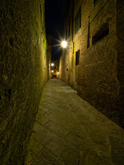 Narrow street in Siena, illuminated by warm streetlights under night sky. Historic architecture, stone-paved path, and serene ambiance create a picturesque nighttime scene. Travel to Tuscany, Italy.