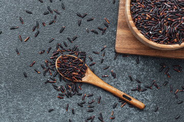 Black rice in wooden bowl with spoon and scattered grains on dark background