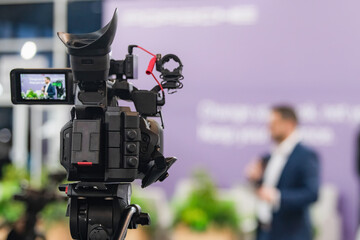 Professional video camera recording a live media conference event, capturing a speaker at the...