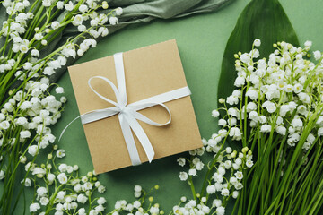 Gift box with white ribbon surrounded by lily of the valley on green background