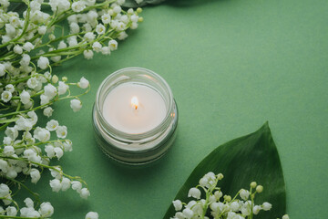 Glass candle burning with white lily of the valley flowers on green background