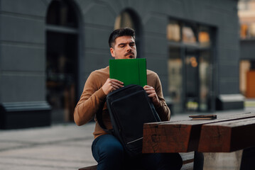 University student putting book in backpack while sitting on bench outdoors