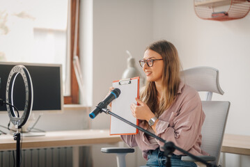 Young woman recording podcast using microphone and clipboard