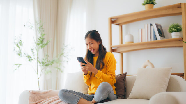 Happy young asian woman relax on comfortable couch at home texting messaging on smartphone, smiling girl use cellphone, chatting online message, shopping online from home