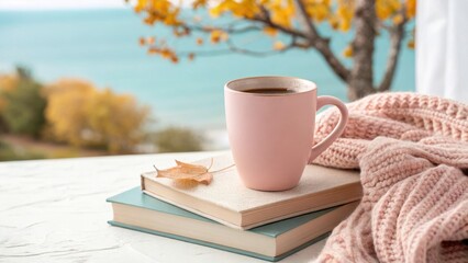 A cozy cup of coffee rests on books with a soft blanket and autumn leaves overlooking a tranquil blue sea