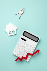 A calculator with a red upward arrow, a house shape, and keys on a ligt teal background, symbolizing rising home value and real estate investment growth. Top view, flat lay.