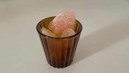 Several pieces of pale pink Himalayan salt crystals fill a brown glass cup, resting on a light -...