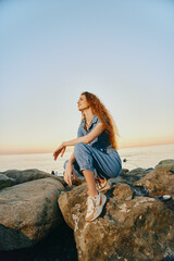 lifestyle woman in modern boho-western denim outfit poses on rocks by the sea during golden hour with warm film color tones and relaxed street style vibe