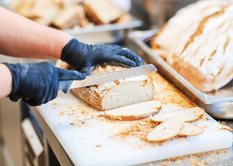 Close-up of a person slicing fresh artisan bread on a cutting board in a professional kitchen. Crumbs, gloves, and bakery atmosphere convey food prep hygiene.