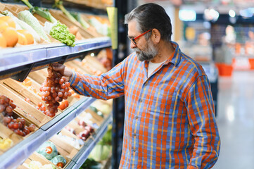 Stylish senior man in red shirt choosing vegetables and fruits at supermarket