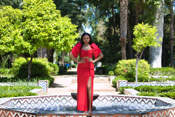 African woman, with long brown hair, young and beautiful dressed in an elegant red dress standing posing on the edge of a golden tiled fountain in seville. The woman is doing different poses.