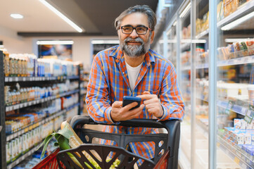Senior Man doing grocery shopping at the supermarket, he is using apps on his smartphone