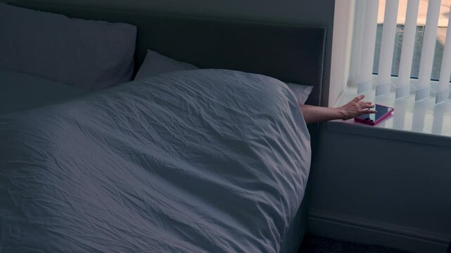 A young woman is sleeping. She is woken up by the sound of the alarm clock. She tries to reach it to turn it off and lie down for just a little longer.