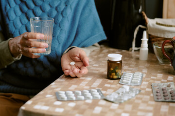 Senior Caucasian woman sitting at table holding pill in one hand and glass of water in other hand, surrounded by various medication blister packs and pill bottle