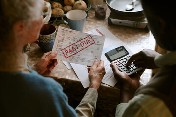 Senior Caucasian woman and senior Black man reviewing overdue utility bill with past due notice,...