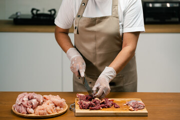 Asian man's hands cutting meat on a wooden cutting board in the kitchen, preparing to cook.