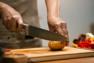 Close-up of Asian man's hands slicing potatoes on a wooden cutting board in the kitchen