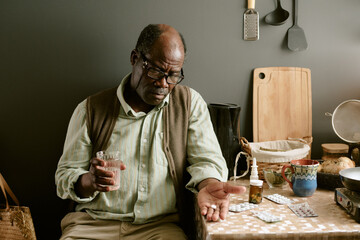 Senior Black man sitting at kitchen table holding glass of water and looking at pills in hand,...