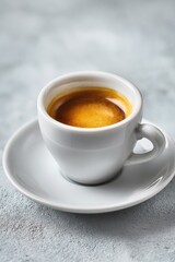 A single espresso shot in a small, white cup and saucer rests on a light gray textured surface.  The crema is rich and golden brown