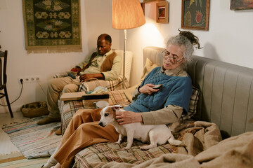 Senior Caucasian woman sitting on sofa petting small dog and holding remote control, senior Black man sitting in background eating meal, both appearing in modest home setting