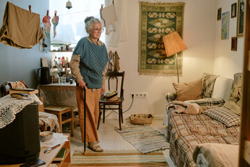Senior Caucasian woman standing with cane in modest living room, wearing glasses and smiling...