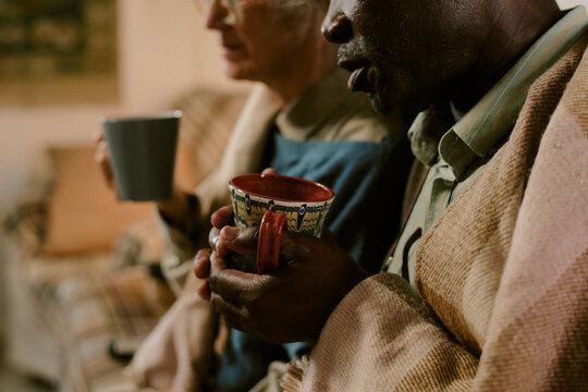 Senior Caucasian woman and senior Black man sitting closely together holding mugs, wrapped in blankets, sharing quiet moment, partially visible faces, focus on hands and cups - Powered by Adobe