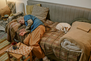 Senior Caucasian woman lying on bed next to small dog, resting with eyes closed, surrounded by...