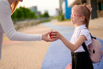 child receiving apple from woman outdoors, symbolizing healthy lifestyle and education. young girl wearing glasses and backpack suggests school setting and nutritious choices.