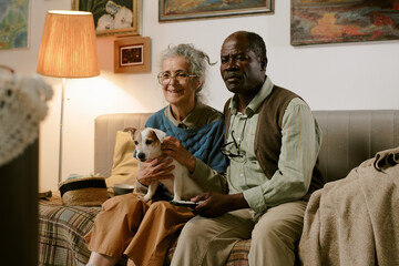 Senior Caucasian woman and senior Black man sitting together on sofa holding small dog, both...