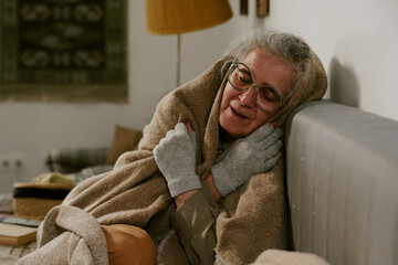Senior Caucasian woman sitting on couch wrapping herself in blanket, wearing glasses and gloves,...
