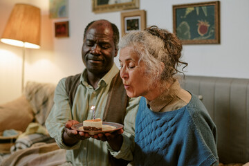 Senior Caucasian woman blowing out candle on small cake while senior Black man holding plate, both...