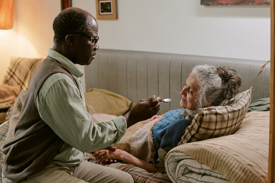 Senior Black man feeding senior Caucasian woman lying on couch, both showing signs of aging, man gently holding spoon near woman's mouth, intimate caregiving moment in modest home - Powered by Adobe
