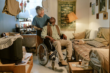 Senior Caucasian woman standing behind senior Black man sitting in wheelchair with disability, both smiling and making eye contact in modest home interior, showing companionship