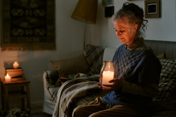 Senior Caucasian woman sitting on sofa holding large candle jar with gentle expression, surrounded...