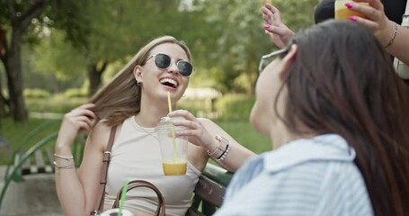 Two carefree friends are enjoying a beautiful summer day outdoors. They are seen laughing and chatting while sipping refreshing drinks on a park bench.