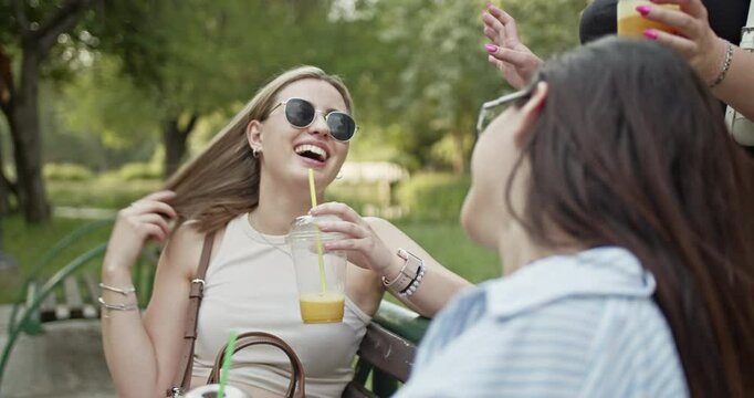 Two carefree friends are enjoying a beautiful summer day outdoors. They are seen laughing and chatting while sipping refreshing drinks on a park bench.