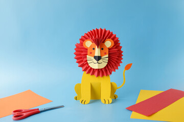 Lion made from colored paper with red folded mane and yellow body placed on blue background, surrounded by scissors and colored sheets. Jungle animal theme for children. Front view with copy space.